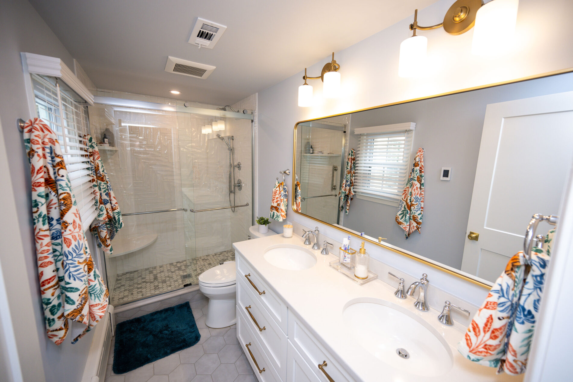 Modern bathroom with dual sinks, large mirror, and glass-enclosed shower. Bright lighting, floral towels, and a teal bath mat accent the white and grey color scheme.