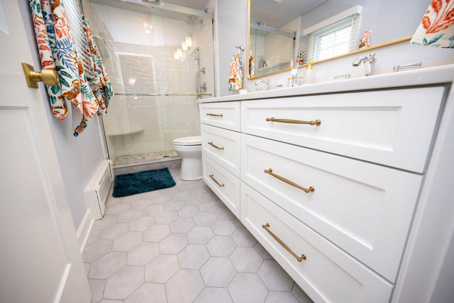 Image by Lesnick Photo Bathroom with white cabinets, hexagonal tile flooring, a toilet, and a glass shower. Colorful floral curtains hang by the window.