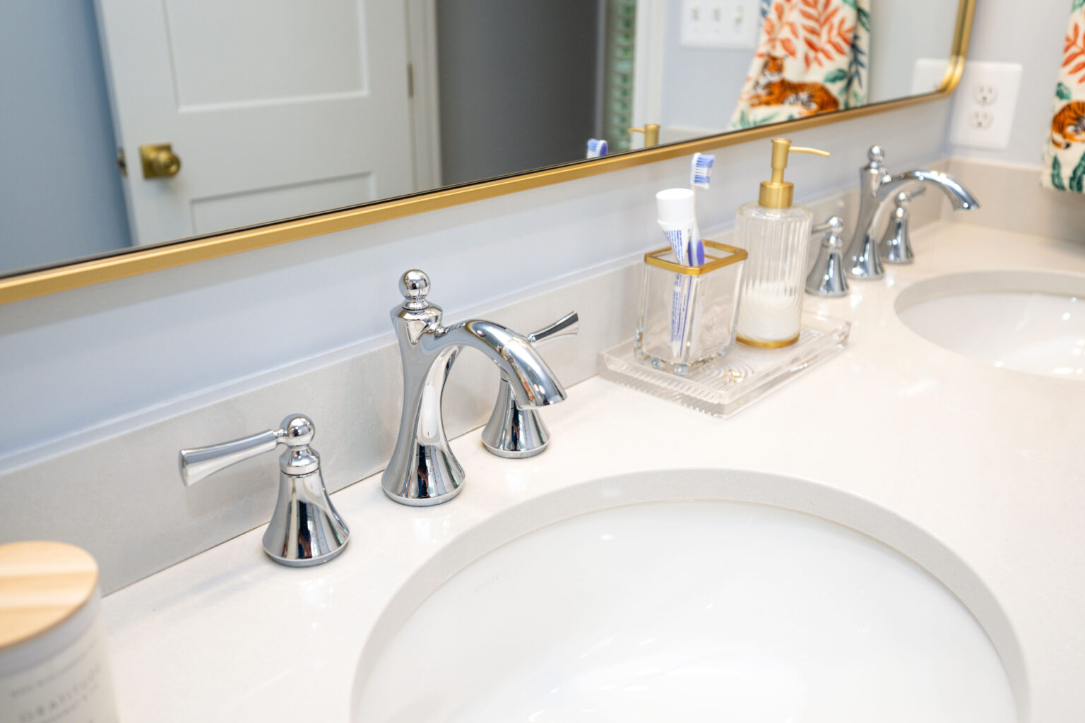 Image by Lesnick Photo A bathroom countertop with a large mirror, featuring dual sinks, chrome faucets, a soap dispenser, and a toothbrush holder. A floral towel is partially visible on the right.