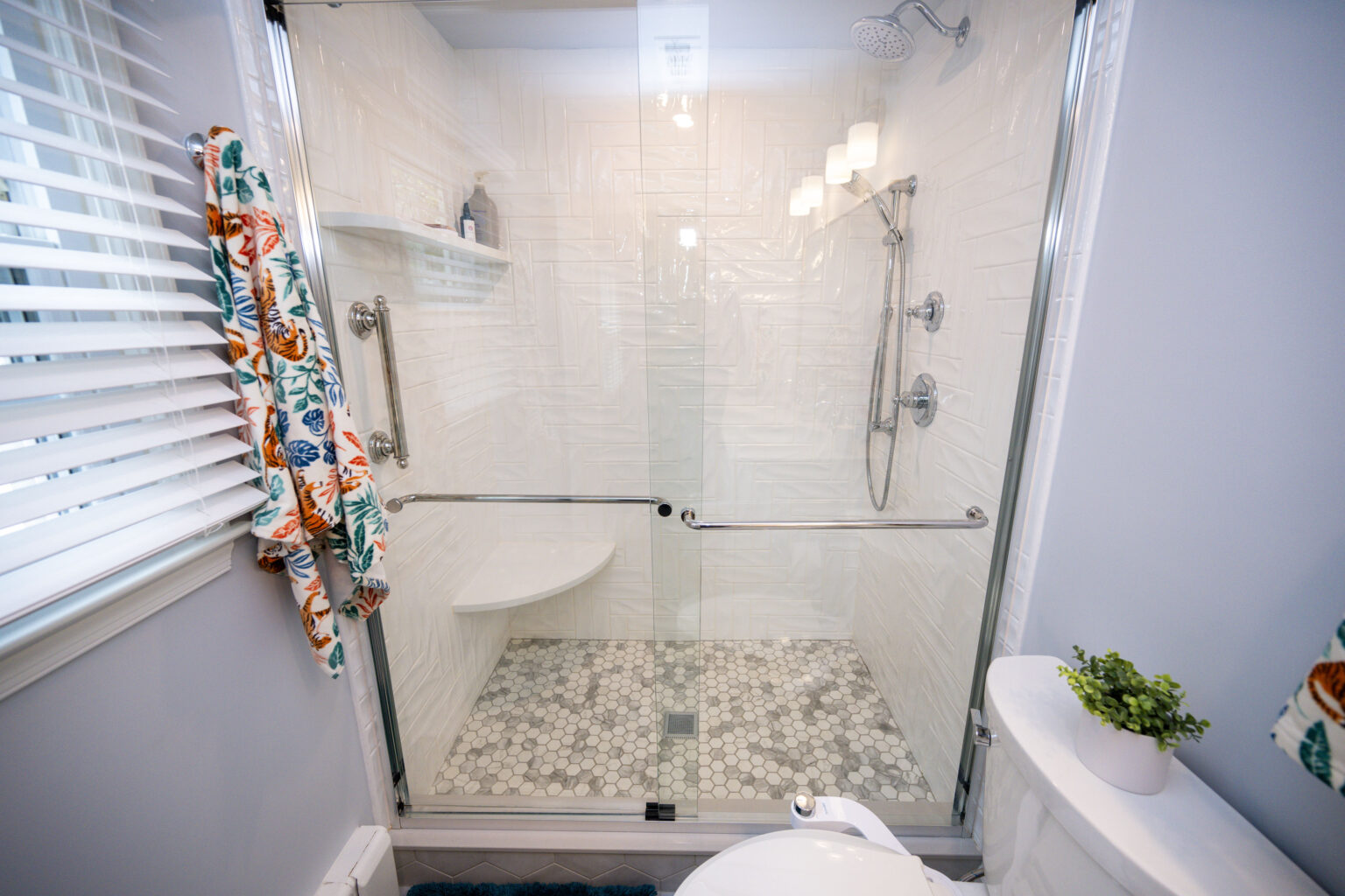 Image by Lesnick Photo Bathroom with a glass-enclosed shower featuring white tile walls, a gray and white mosaic floor, and a wall-mounted showerhead. A colorful towel hangs nearby, and a plant sits on the toilet tank.