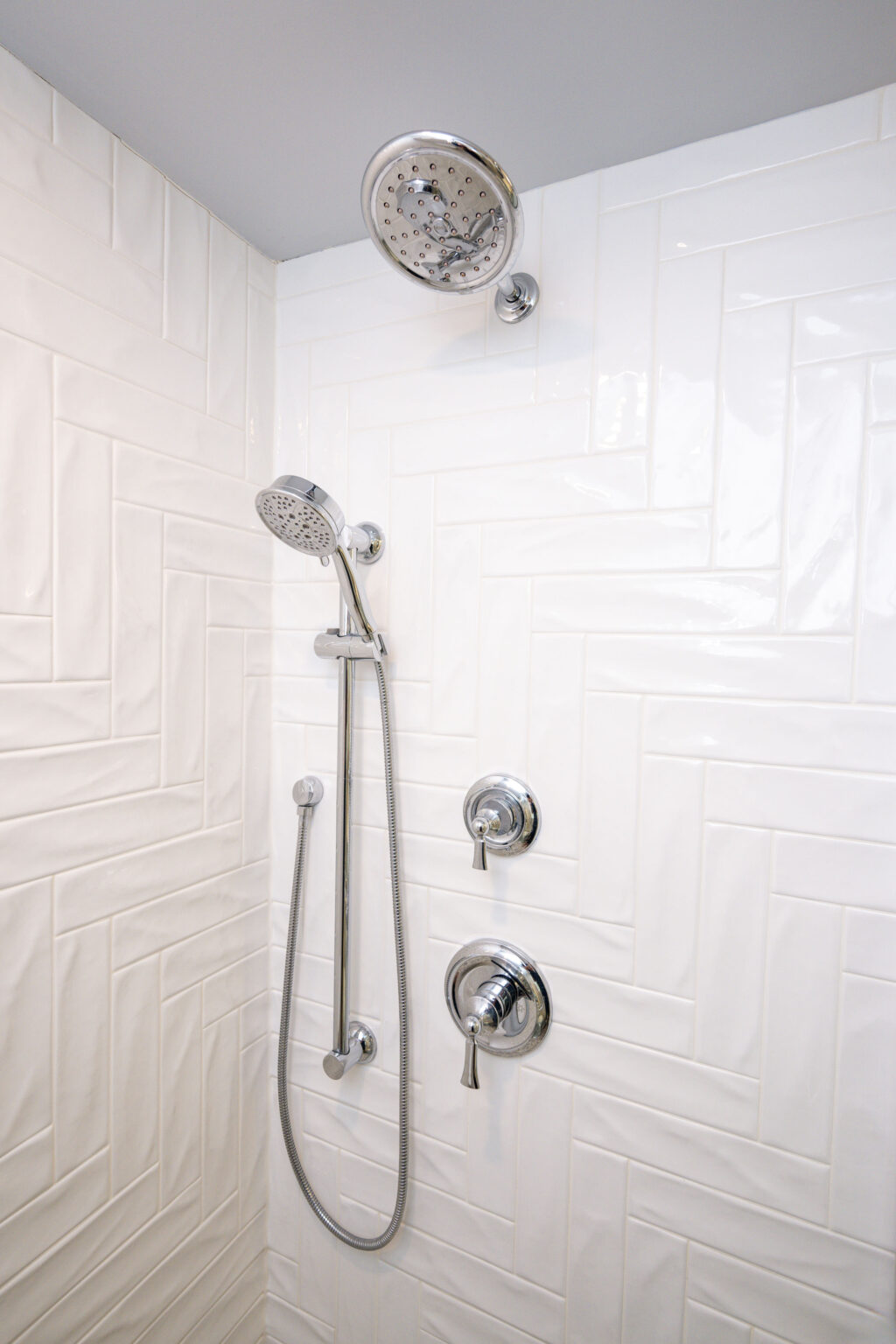 Image by Lesnick Photo White tiled shower with two chrome showerheads, one fixed and one handheld, and matching controls.