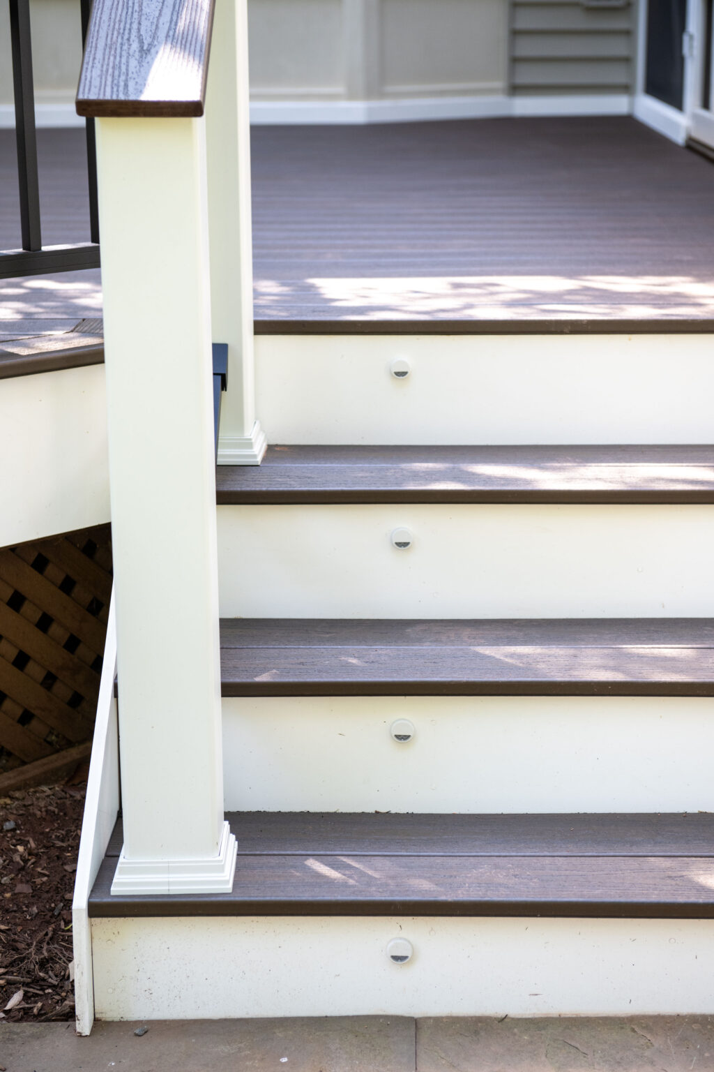 Image by Lesnick Photo Outdoor stairs with four steps leading to a deck, featuring dark brown treads and light-colored risers, with small round lights embedded in each riser.