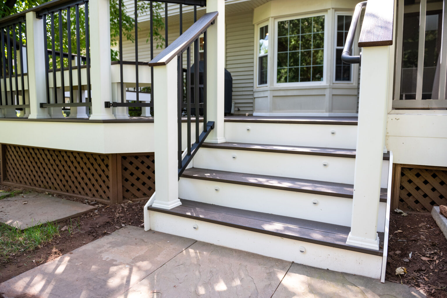 Image by Lesnick Photo Wooden deck stairs with white railings and dark handrails lead to a house. The base features latticework, and stone slabs form a pathway below.