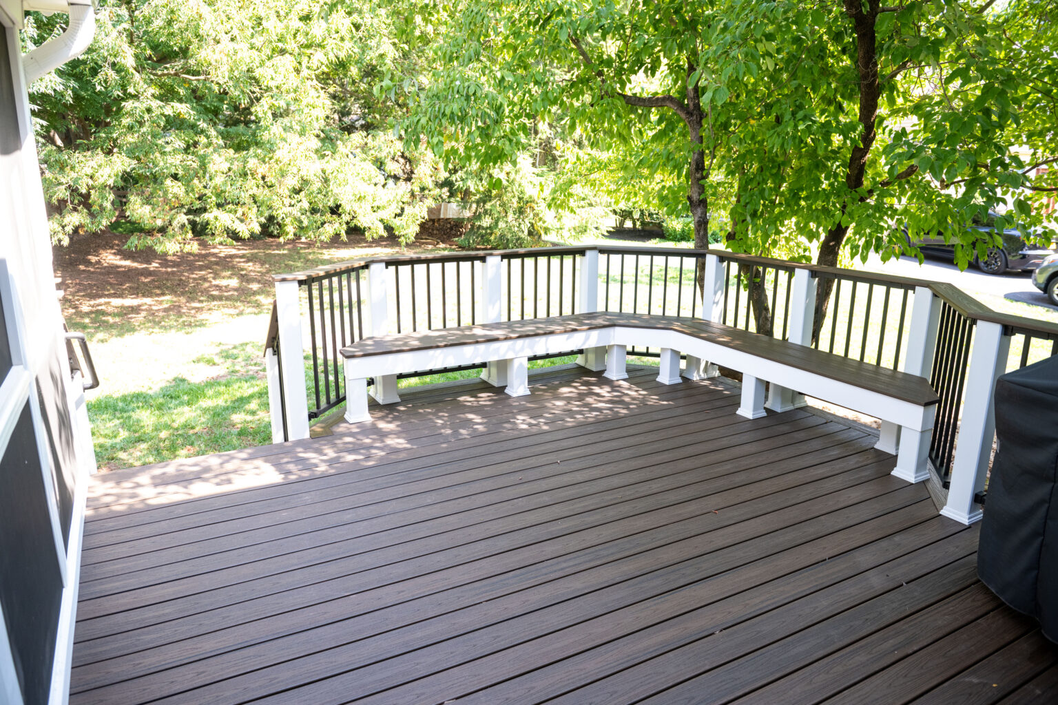 Image by Lesnick Photo Wooden deck with built-in bench seating, surrounded by a black and white railing. Trees and grass are visible in the background.