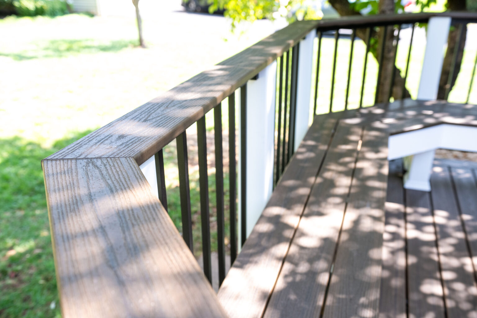 Image by Lesnick Photo Close-up of a wooden deck railing with black balusters, casting tree shadows on the deck. Green grass and a tree are visible in the blurred background.