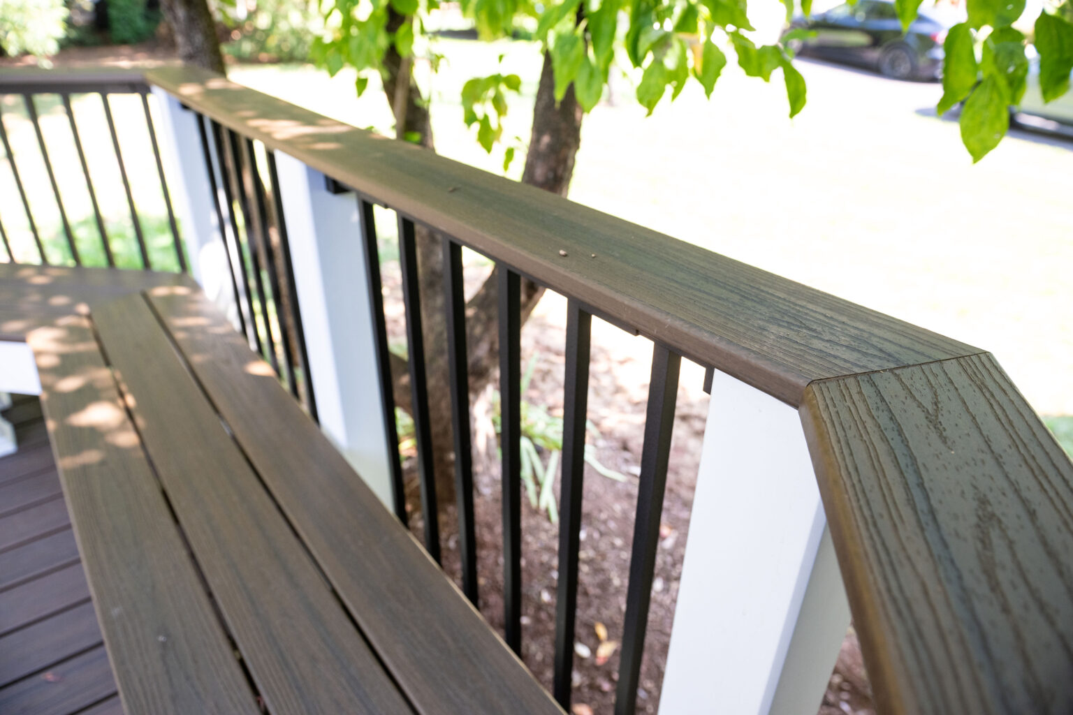 Image by Lesnick Photo A wooden deck railing with vertical black metal balusters is shown, accompanied by a bench-like wooden feature, against a backdrop of trees and grass.