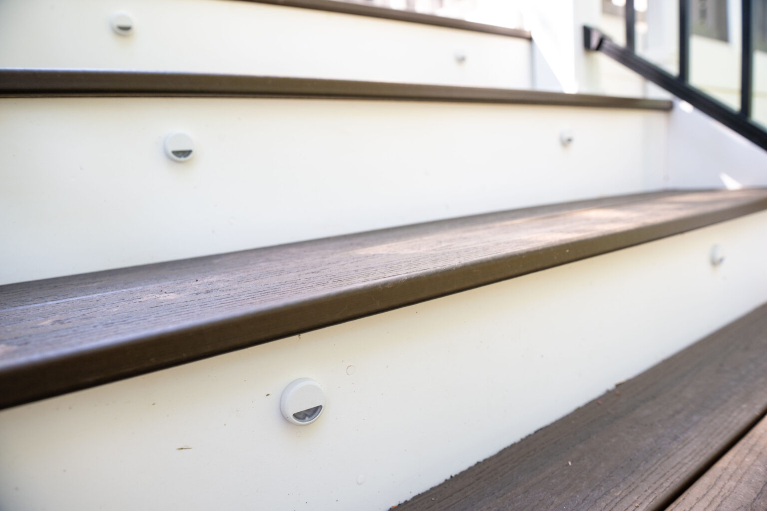 Image by Lesnick Photo Close-up of outdoor wooden steps with white risers and small round lights, leading upwards with a black metal handrail visible on the right.