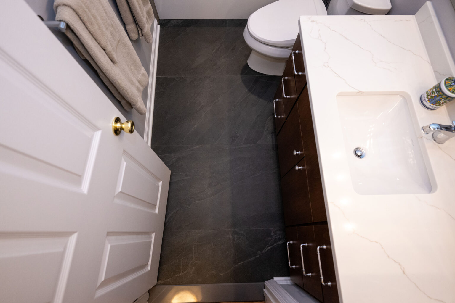 Image by Lesnick Photo Overhead view of a small bathroom featuring a white door, a dark floor, a white countertop with a sink, and a toilet.