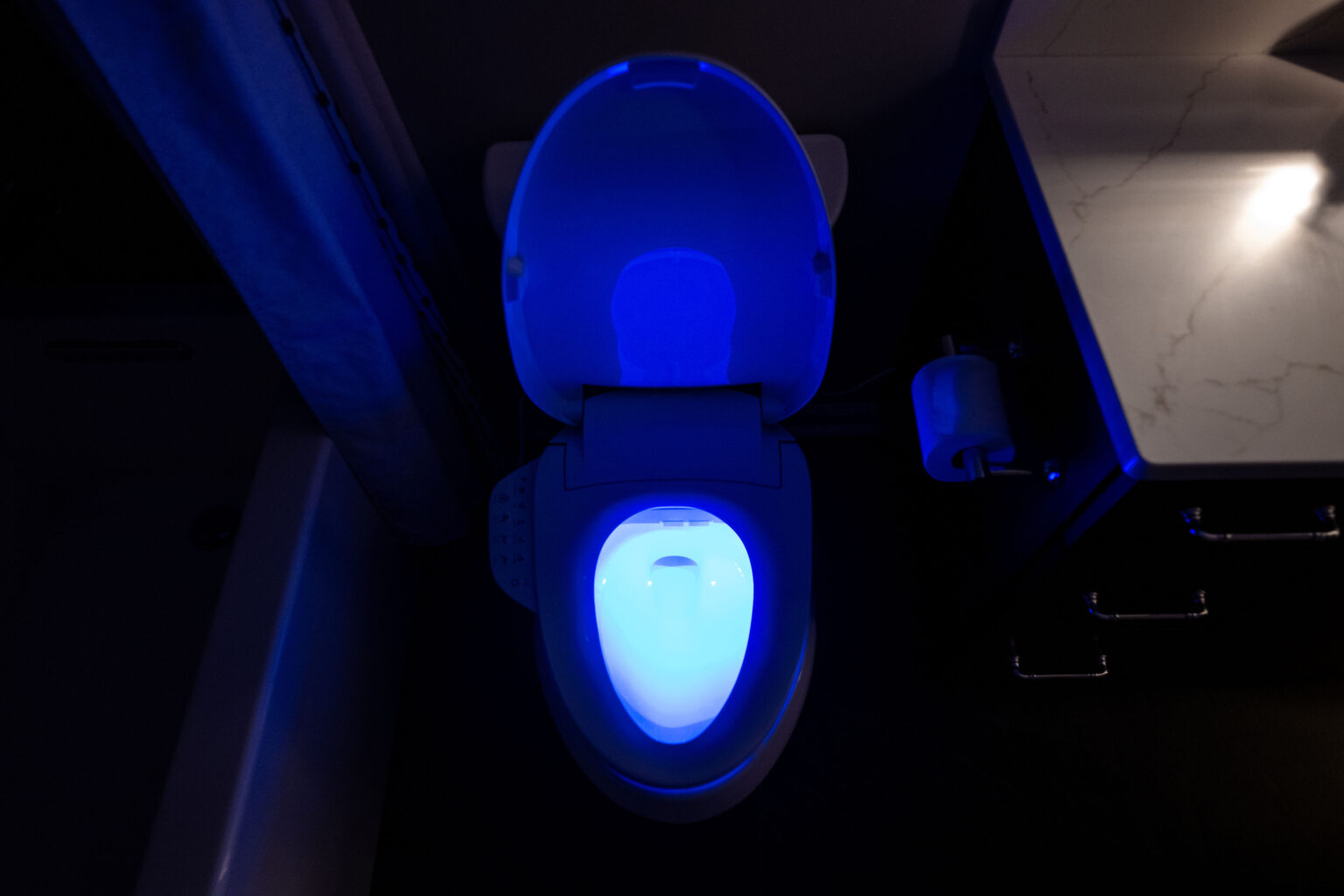 Image by Lesnick Photo A toilet glows with blue LED light in a dimly lit bathroom, next to a vanity with a paper roll on the wall.
