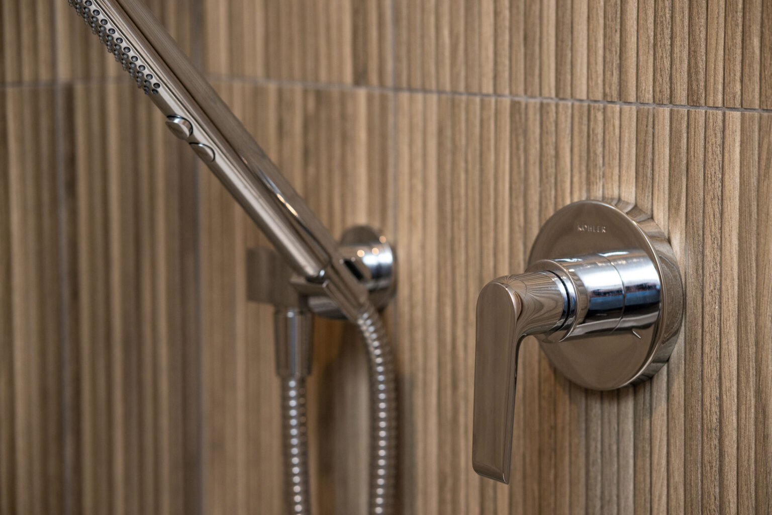 Image by Lesnick Photo A modern shower fixture with a chrome handle and detachable showerhead mounted on textured beige tiles.