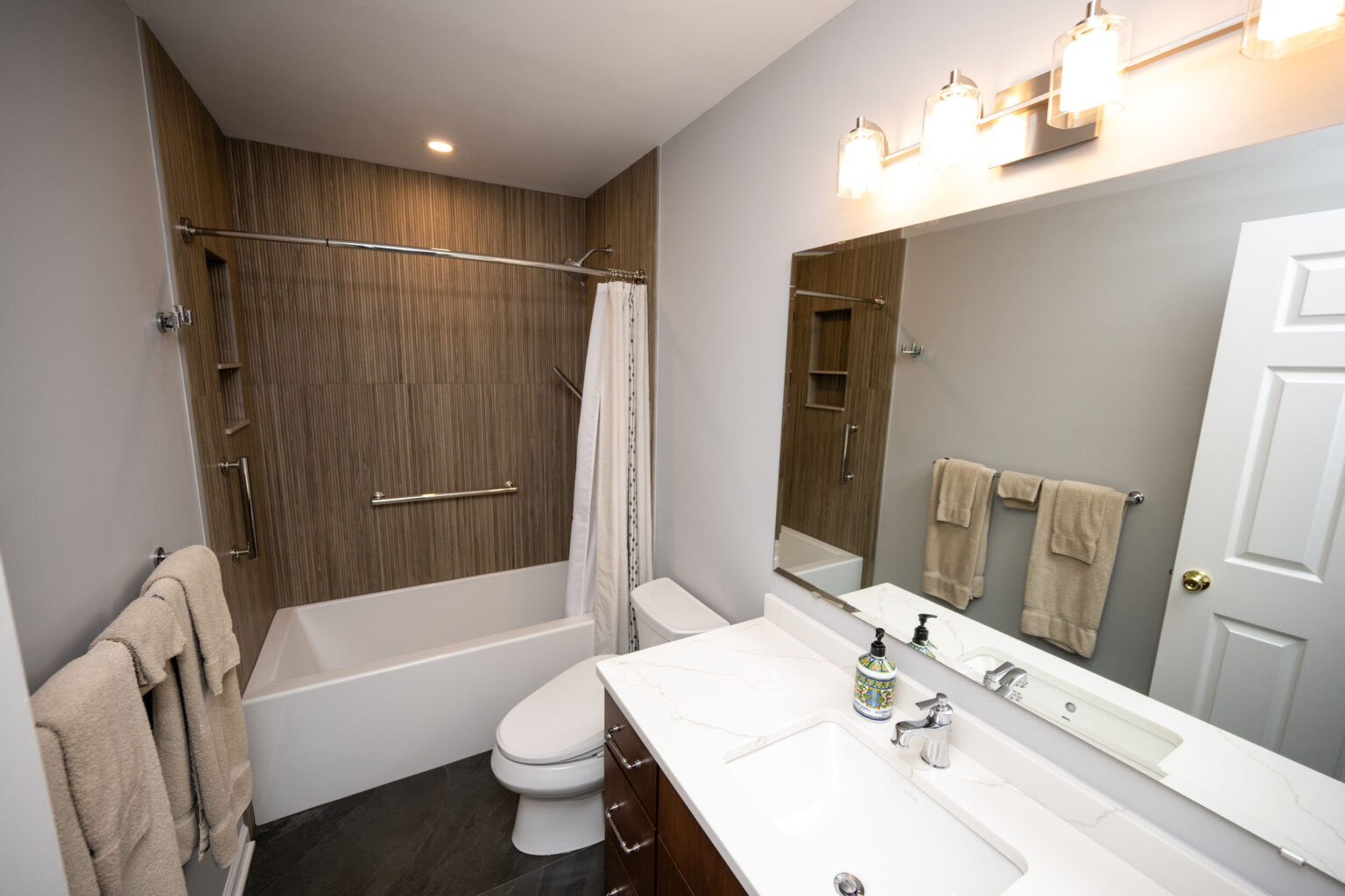 Image by Lesnick Photo Modern bathroom with a bathtub, shower curtain, and wood-paneled wall. A toilet is next to the tub. Double vanity with a large mirror and three-bulb light fixture above. Towels hang nearby.
