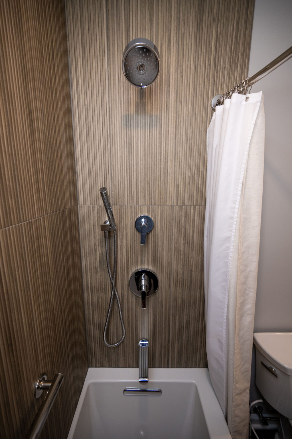 Image by Lesnick Photo A modern shower setup with a handheld showerhead, fixed showerhead, faucet controls, and a white curtain on a curved rod. Brown, striped wall tiles and a white bathtub are also visible.