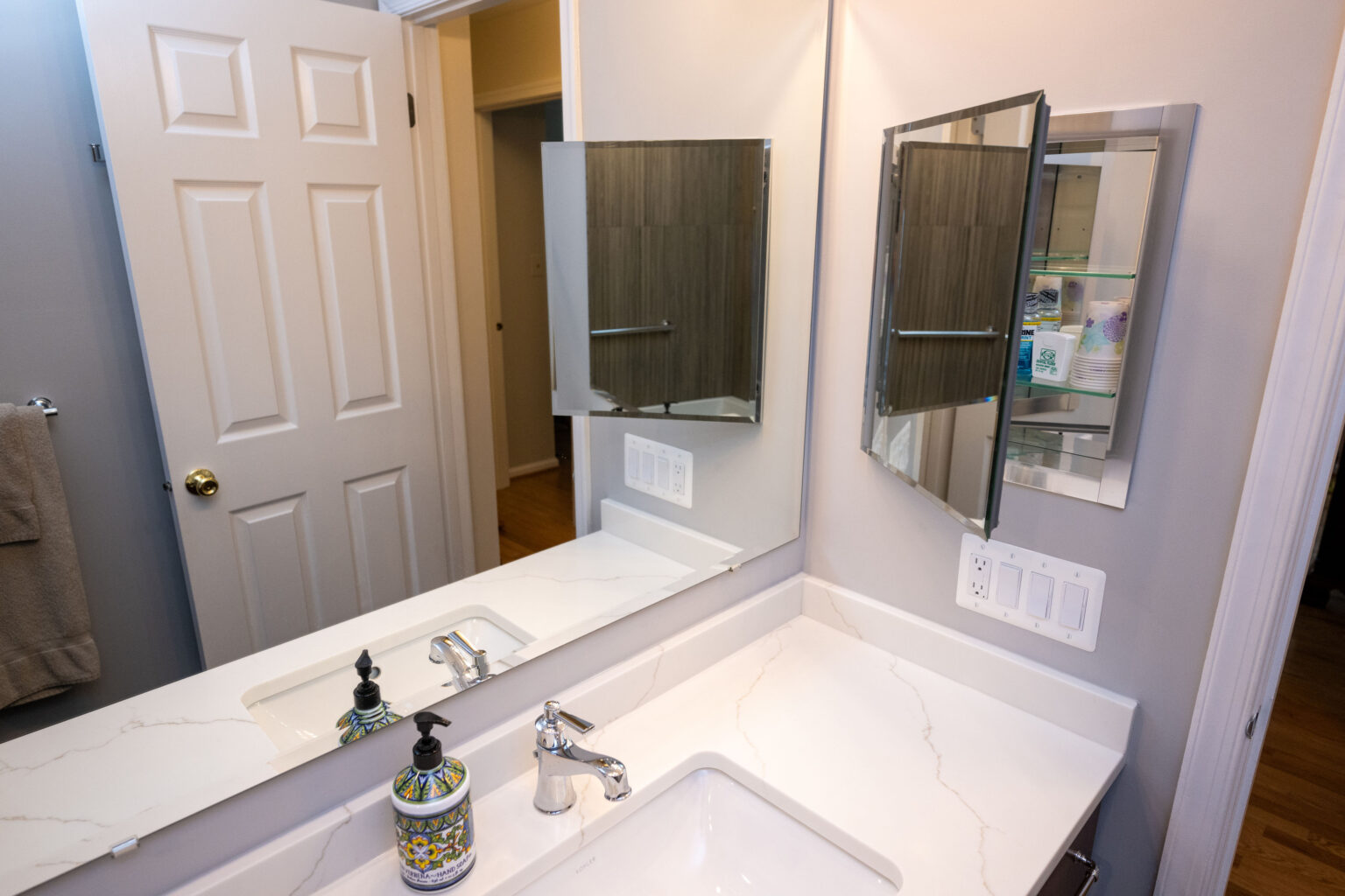 Image by Lesnick Photo Bathroom with white countertop, sink, soap dispenser, and a mirrored medicine cabinet. Door slightly open. Light switches visible on the wall.