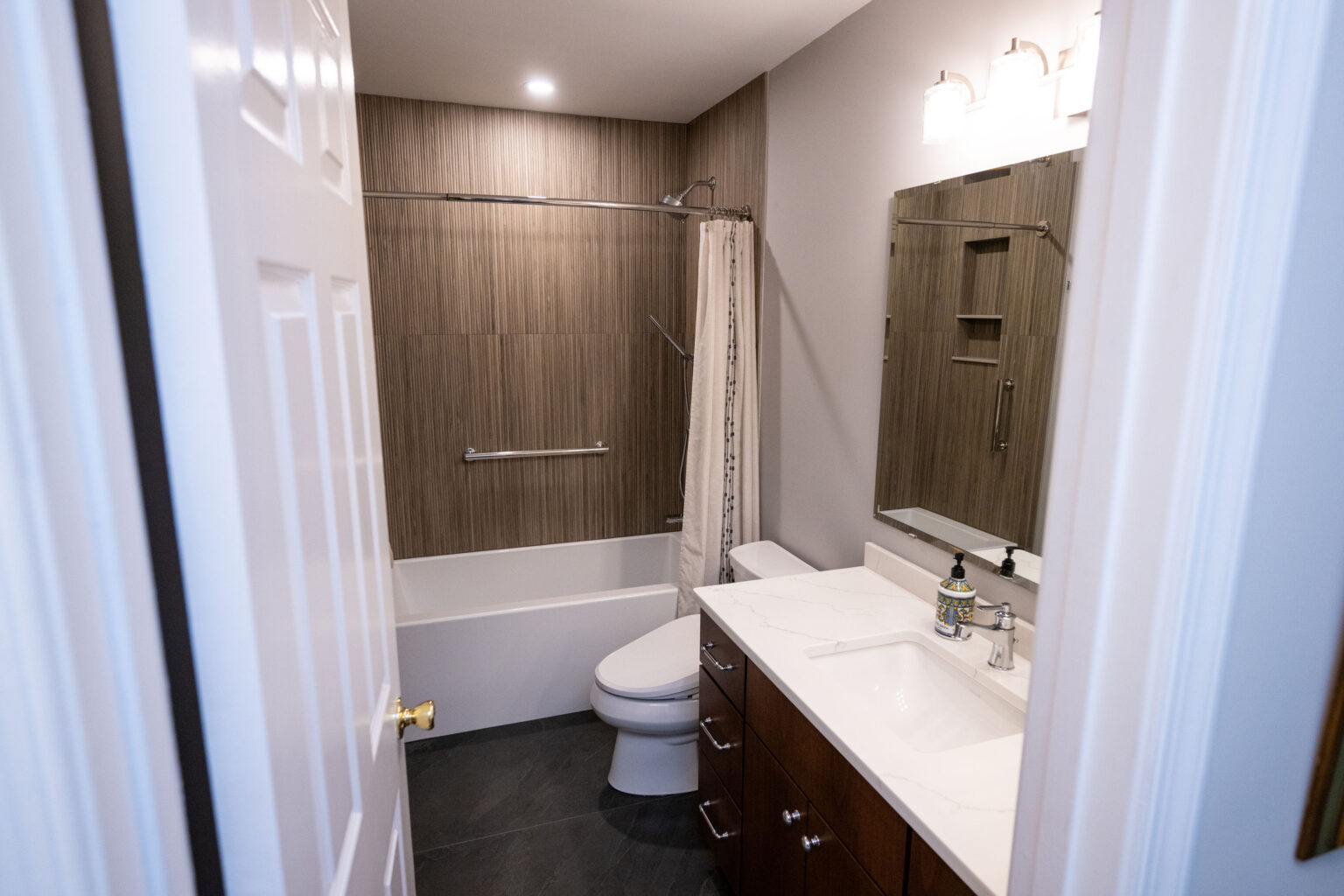 Image by Lesnick Photo Modern bathroom with a white bathtub, brown tiled walls, white sink on a dark wood vanity, large mirror, and a toilet.
