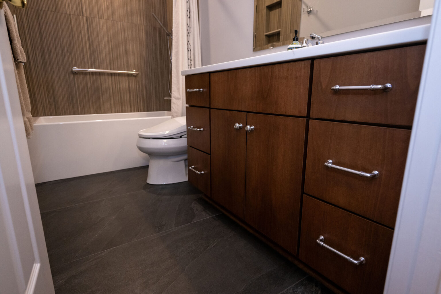Image by Lesnick Photo A modern bathroom with dark wood cabinetry, a white countertop, a toilet next to a bathtub with a wall-mounted grab bar, and dark tiled flooring.