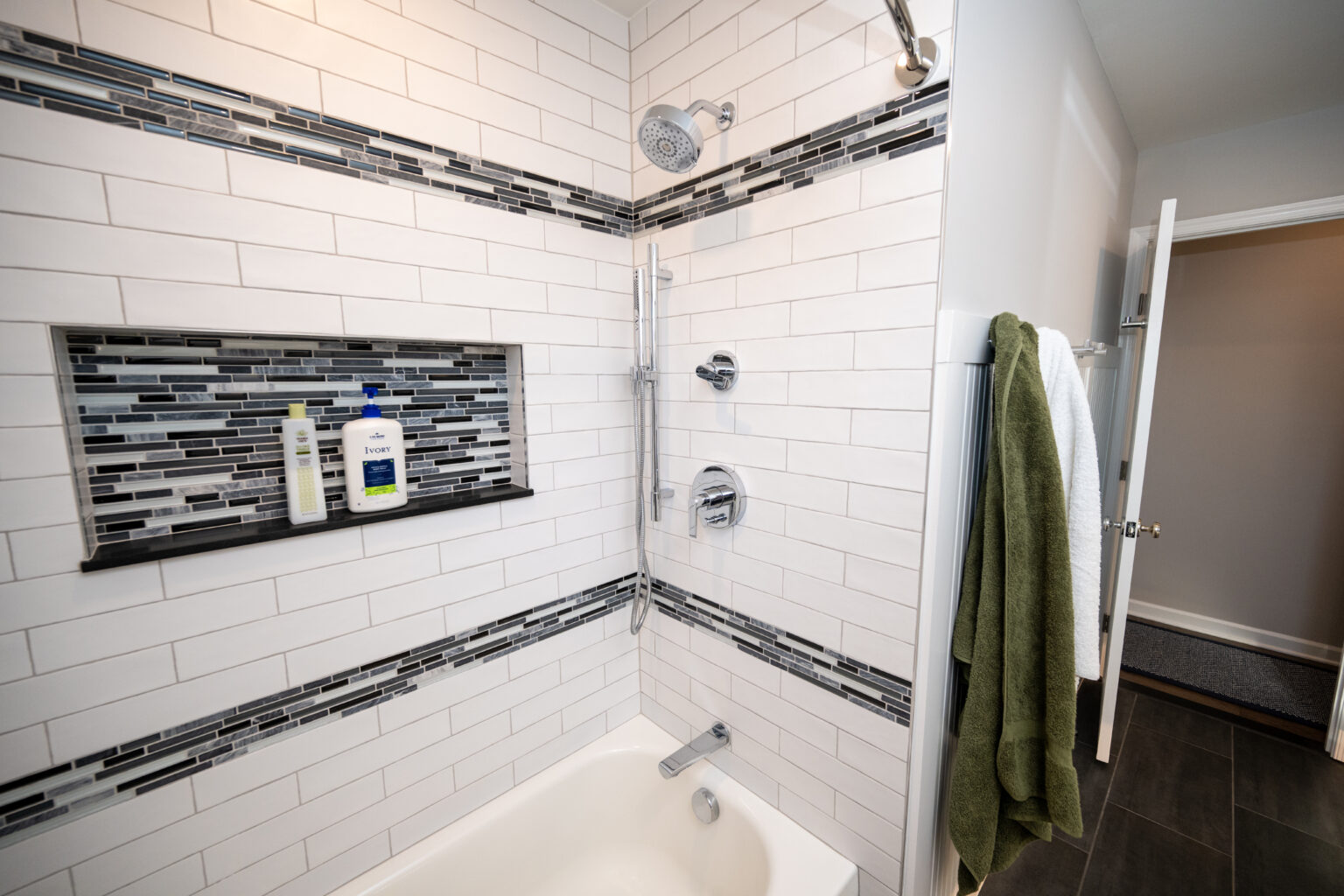 White tiled bathtub and shower with black and gray accent tiles, built-in shelf holding toiletries, and towels hanging on a nearby rack and door.