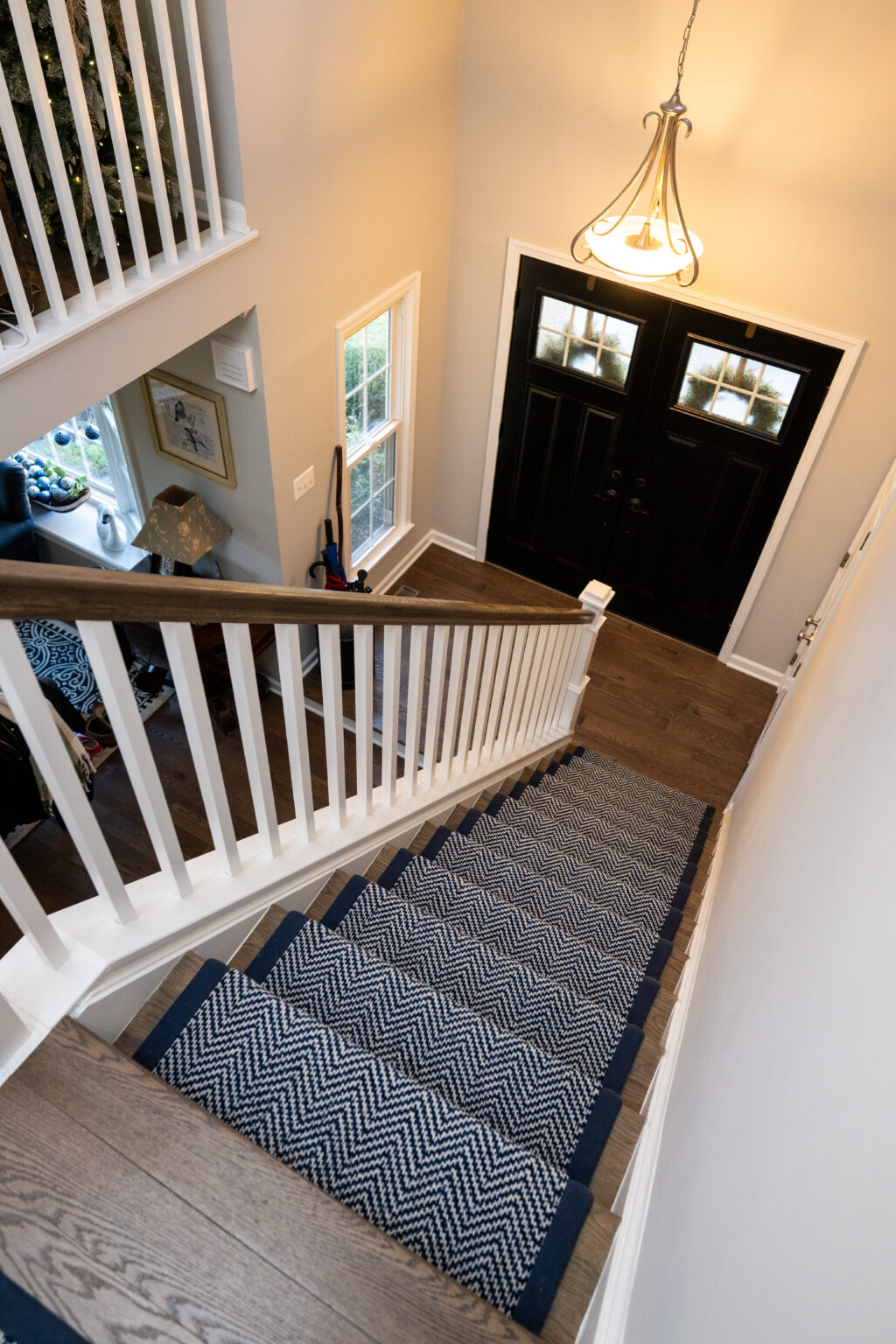 View from the top of a staircase with a herringbone-patterned runner, leading to double black front doors in a well-lit entryway.