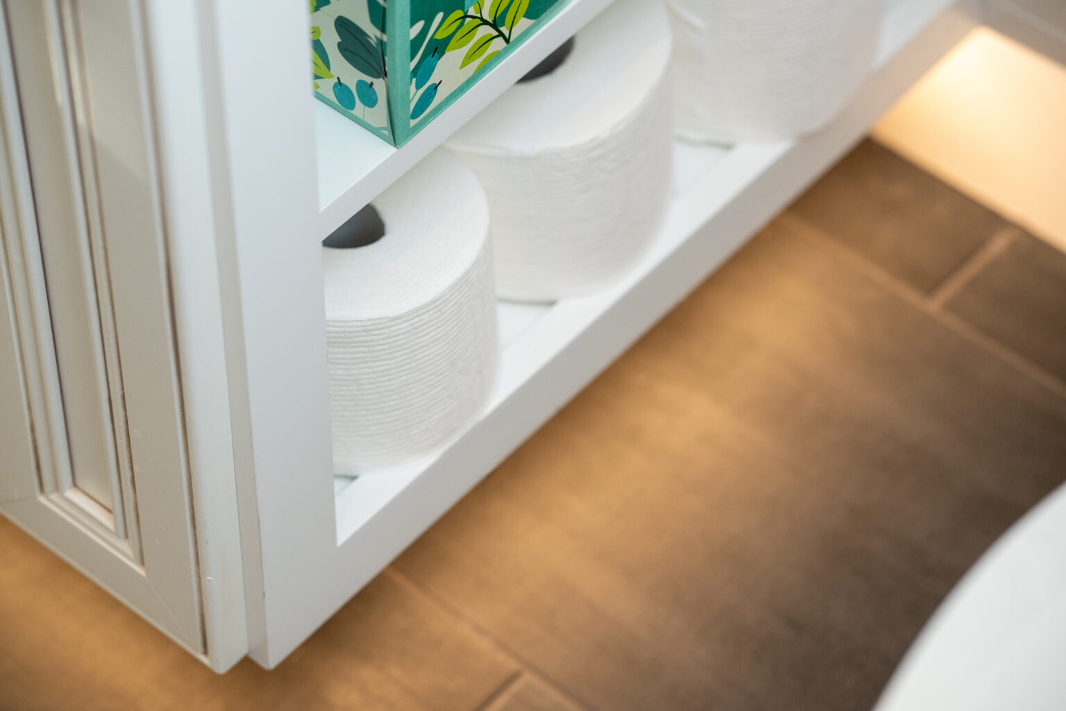 Several rolls of toilet paper are neatly stacked on white shelves next to a patterned tissue box above a tiled bathroom floor.
