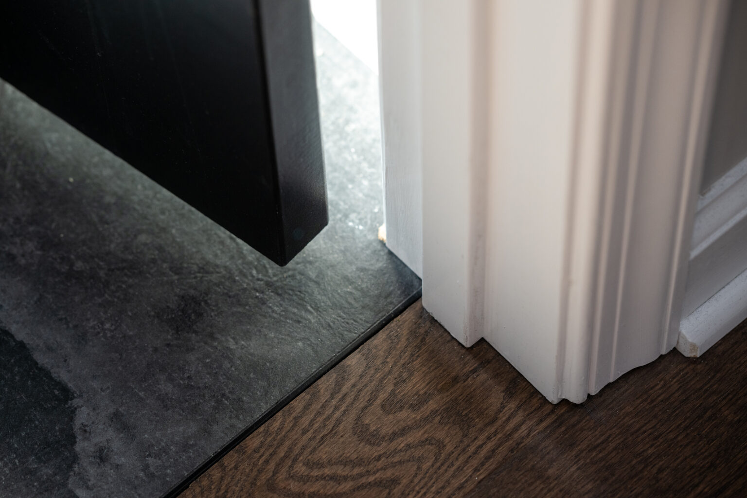 A close-up of a black door slightly open, revealing a transition between gray tile flooring and dark brown wood flooring near a white door frame.