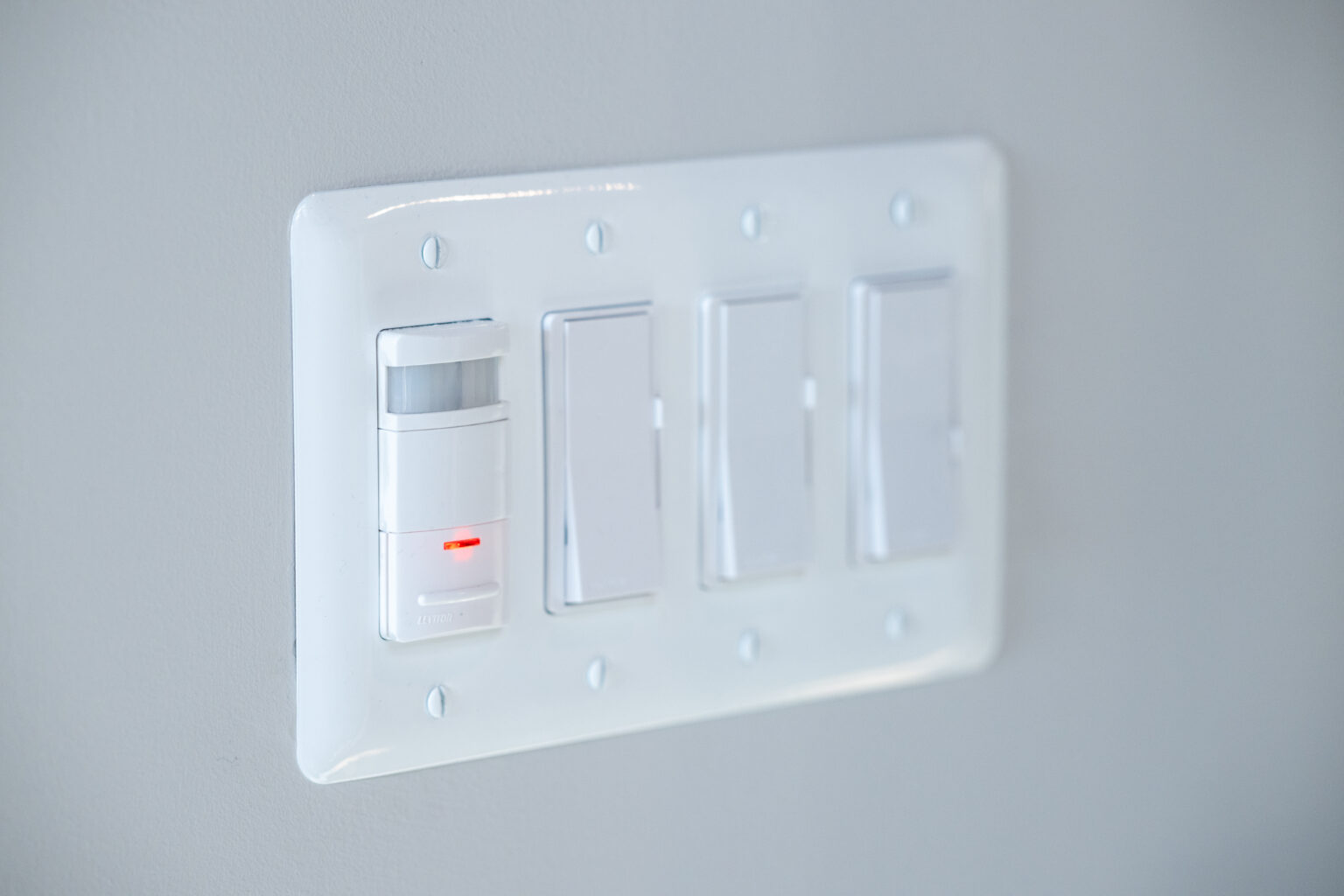 A close-up of a wall panel with four light switches; the leftmost switch has a motion sensor and a small red indicator light.