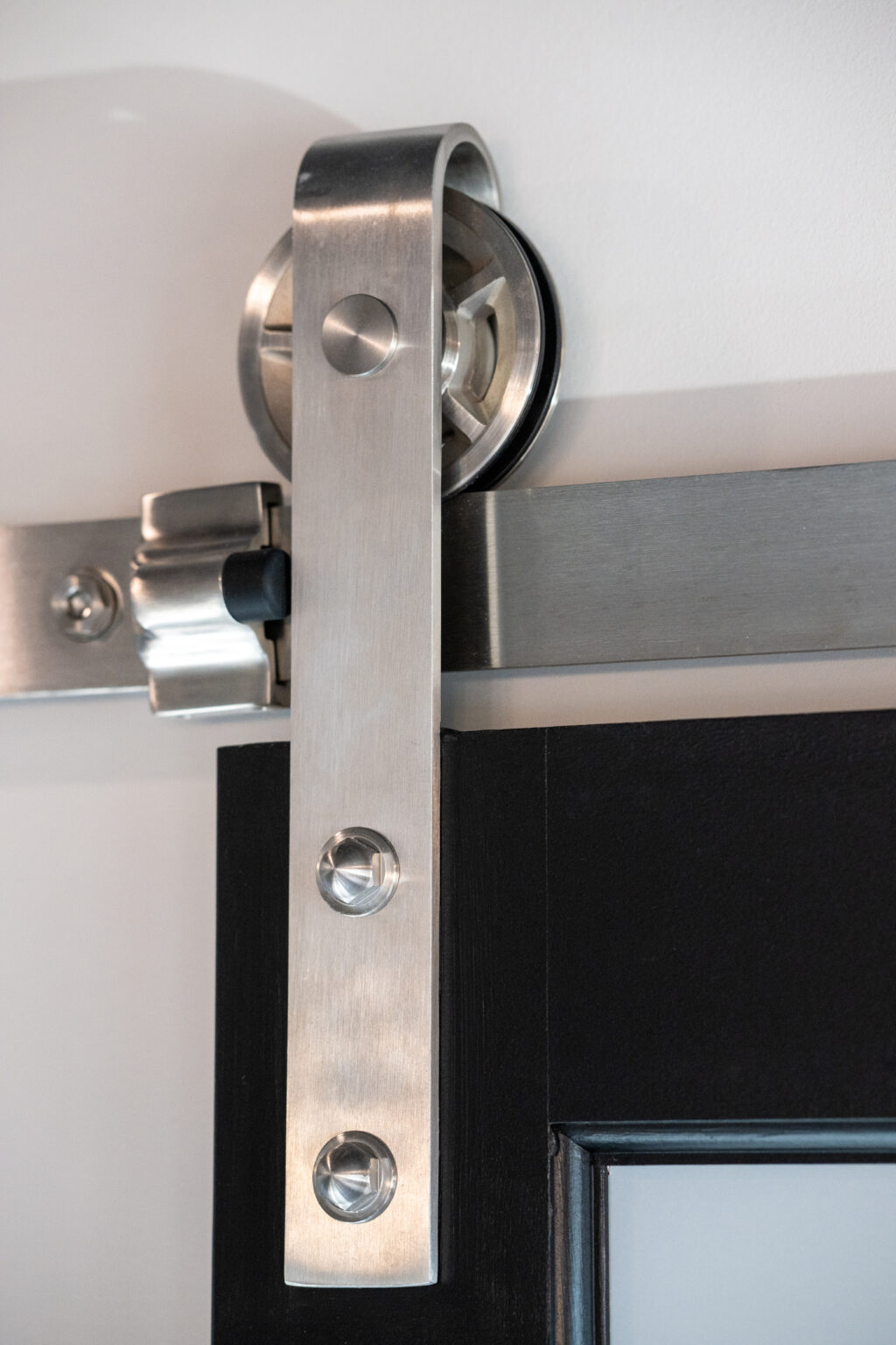 Close-up of a stainless steel sliding barn door hardware track and roller mounted on a white wall and attached to a dark-colored door.