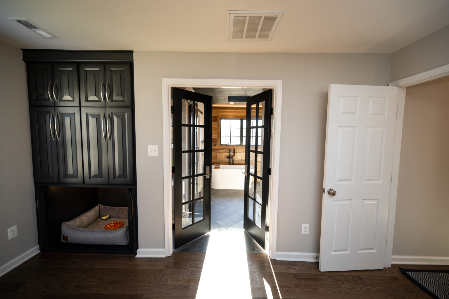 A room with dark wood cabinets, a dog bed, and open French doors leading to a bathroom with a bathtub and window.