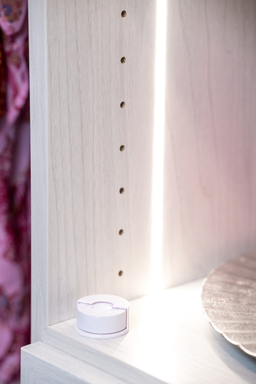 A white plastic device sits on a light wood shelf with adjustable peg holes, next to a decorative metallic plate.