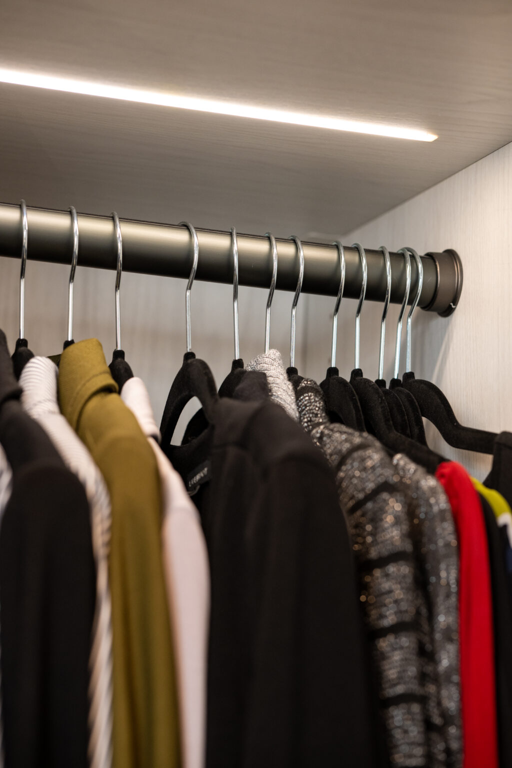 Clothes hanging on metal hangers in a closet, arranged in a row with various colors and fabrics visible under a strip of overhead lighting.