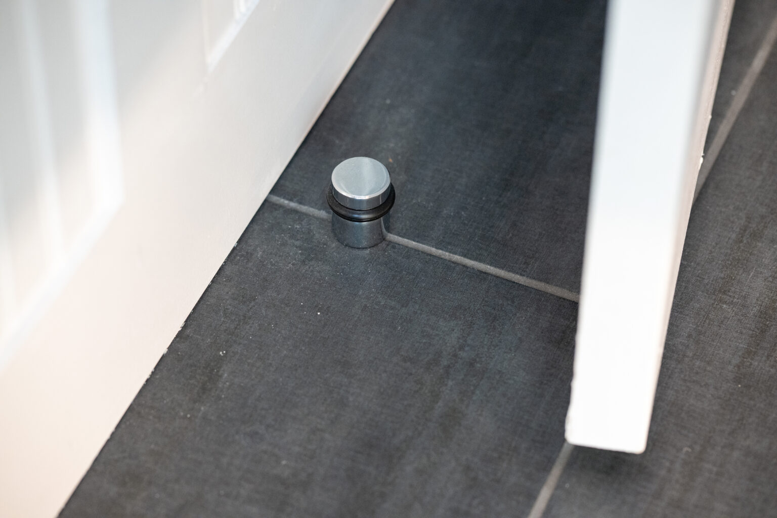 A metal door stopper is installed on a tiled floor, positioned to prevent a white door from hitting the wall.