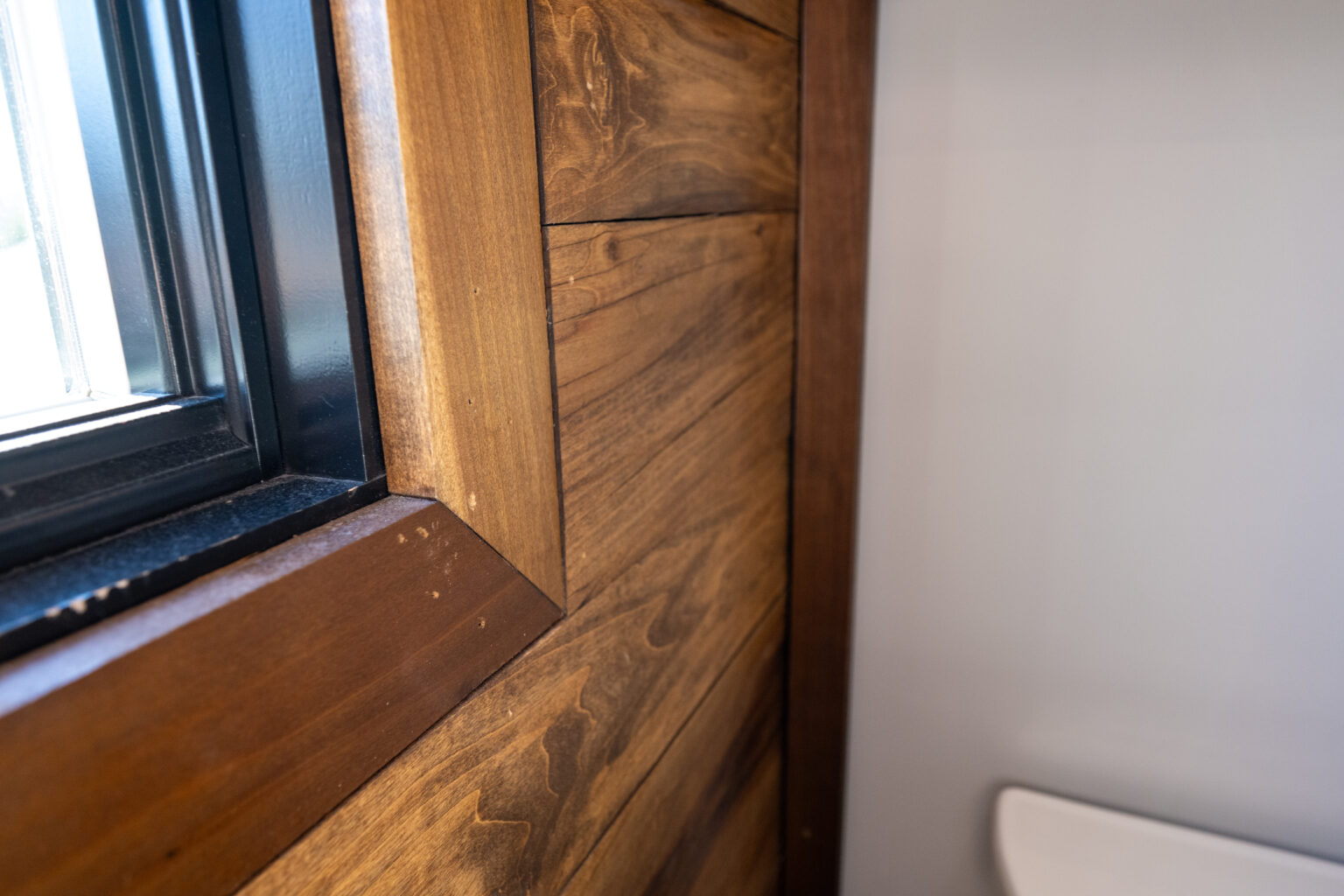 Close-up of a window corner with wooden trim and paneling next to a light gray wall; a partial view of a white fixture is visible on the right.