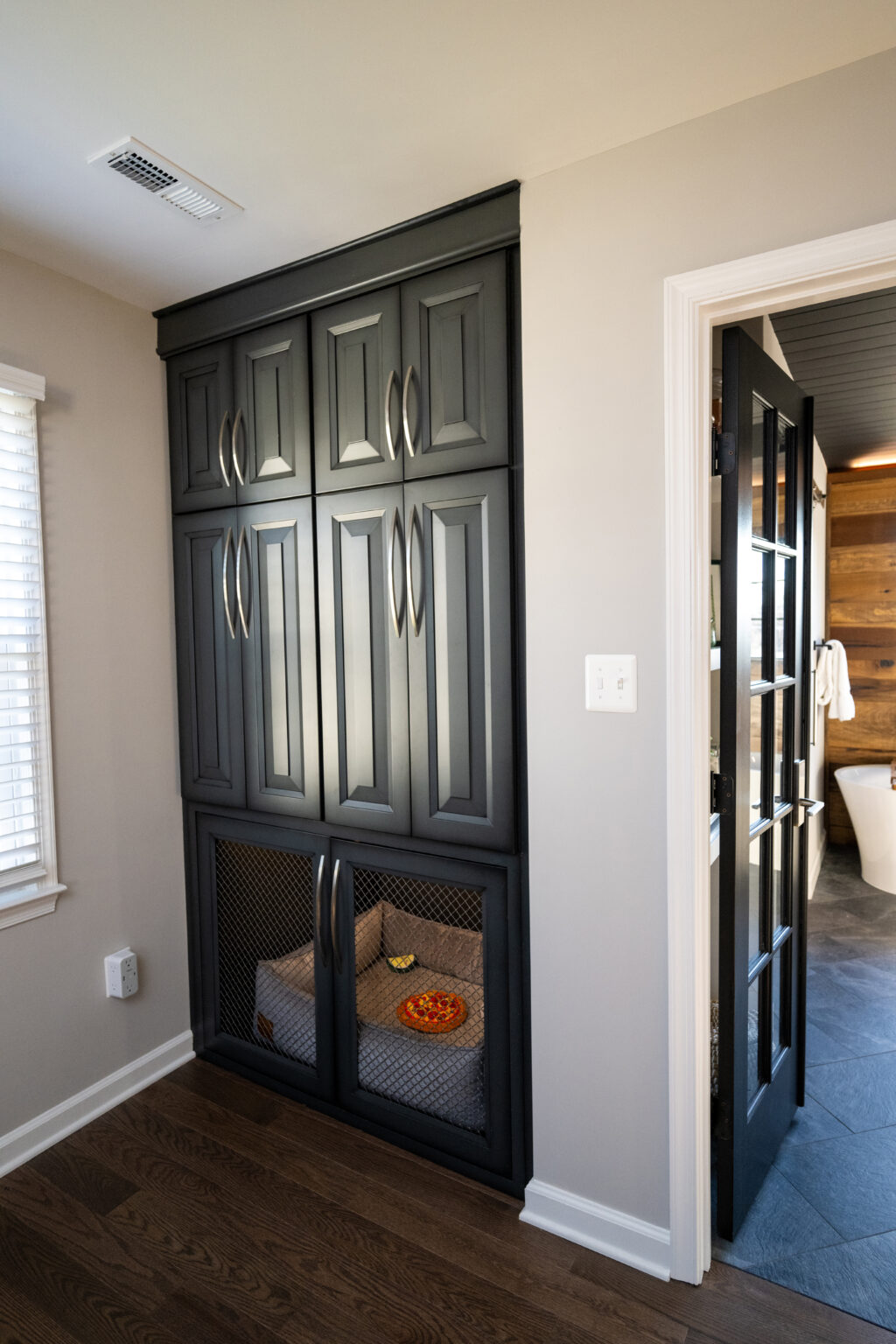 Black built-in cabinet with lower section converted into a dog bed area, featuring mesh doors and a dog bowl inside, in a hallway next to a bathroom.