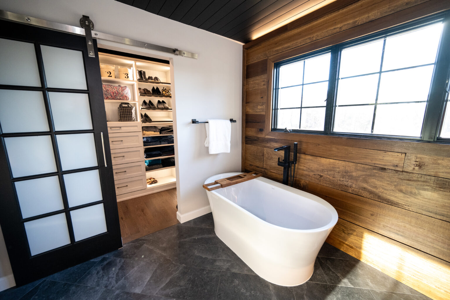 Modern bathroom with a freestanding tub, wooden accent wall, large window, towel rack, and an open sliding door revealing a walk-in closet with shelves and drawers.