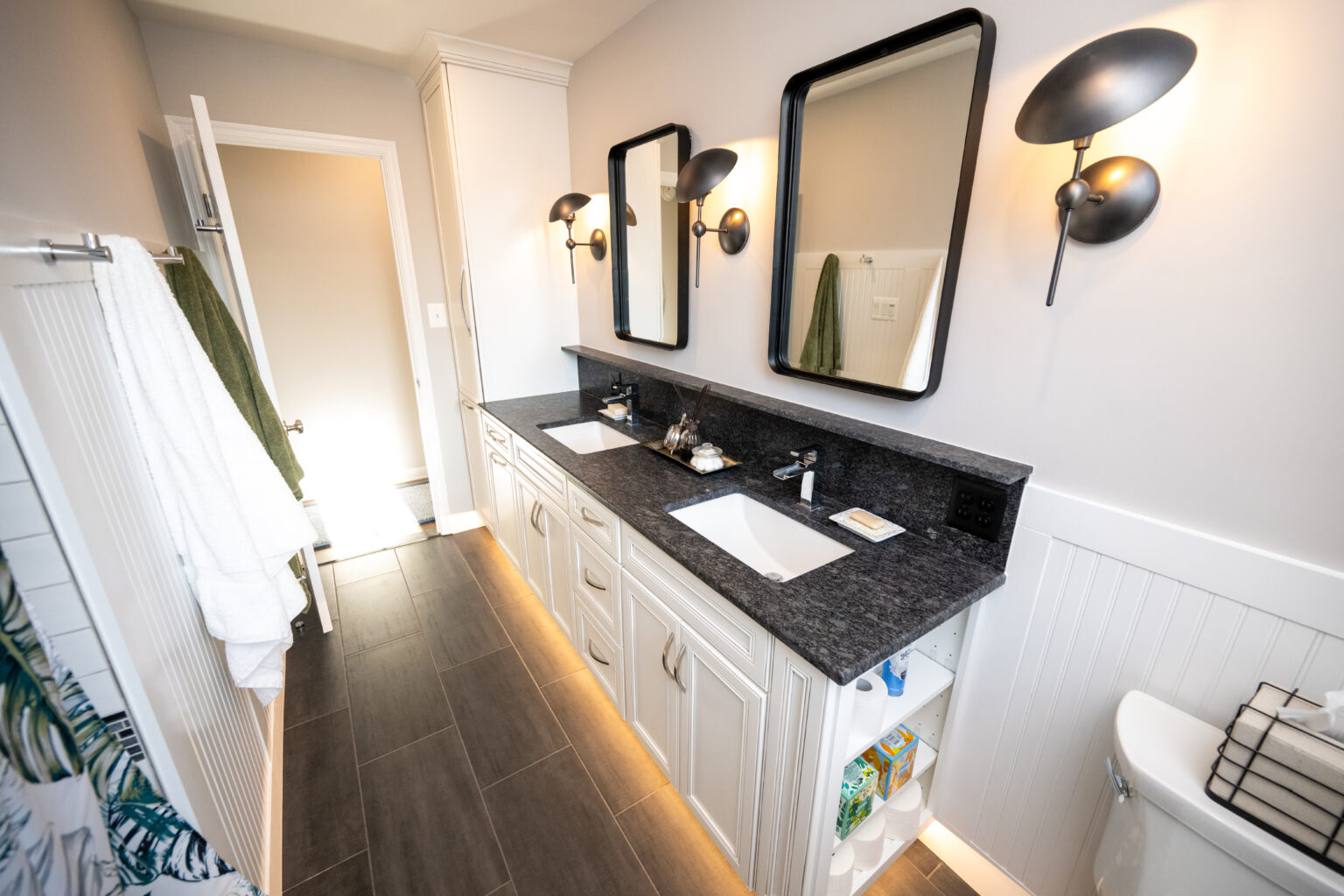 Modern bathroom with double sinks, black countertops, two mirrors, wall-mounted lights, towels on racks, and storage shelves below the sinks.