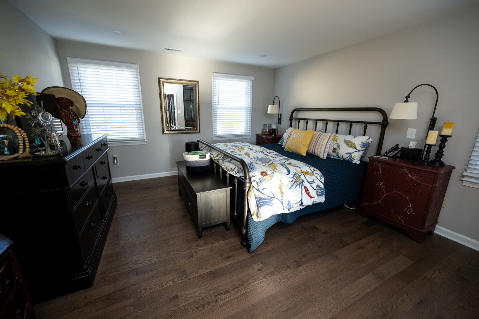 A tidy bedroom with a metal-framed bed, two nightstands, a dresser, a mirror, and hardwood flooring, lit by natural light from two windows.