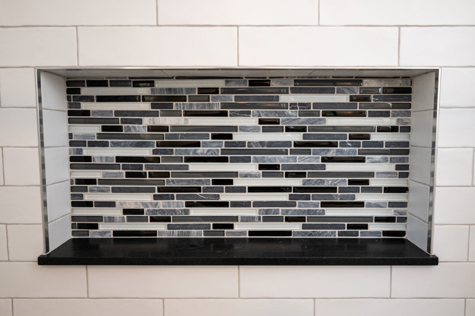 Rectangular alcove with a black shelf and a decorative backsplash of horizontal black, gray, and white tiles, set in a wall of larger white rectangular tiles.