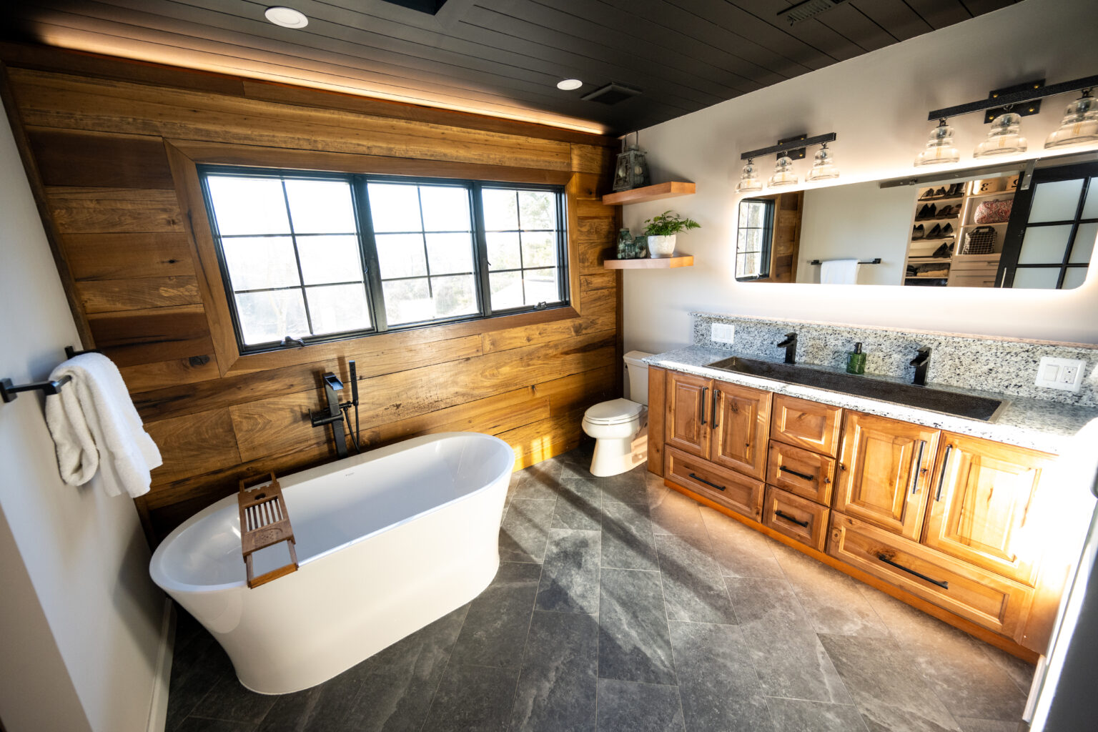 Modern bathroom with a freestanding tub, wooden walls, double sink vanity, large mirror, shelves, and a window allowing natural light into the room.
