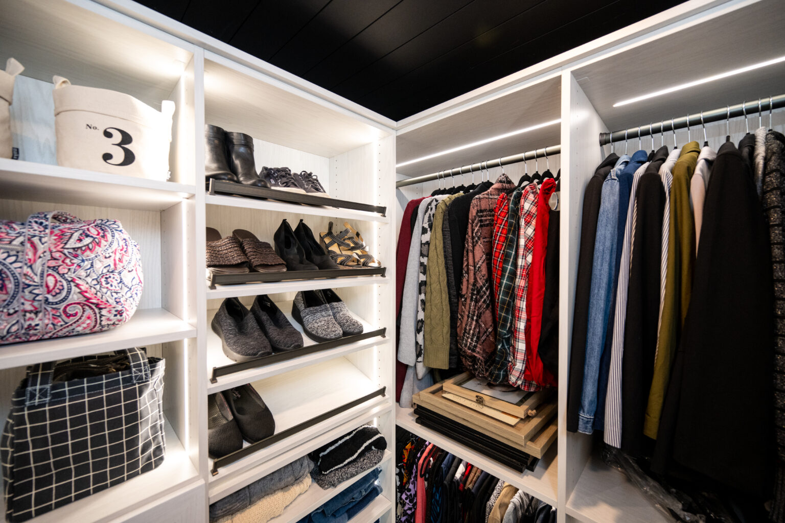 A neatly organized closet with shelves of shoes, folded clothes, storage bins, and a row of shirts, jackets, and plaid tops hanging on a clothes rod.
