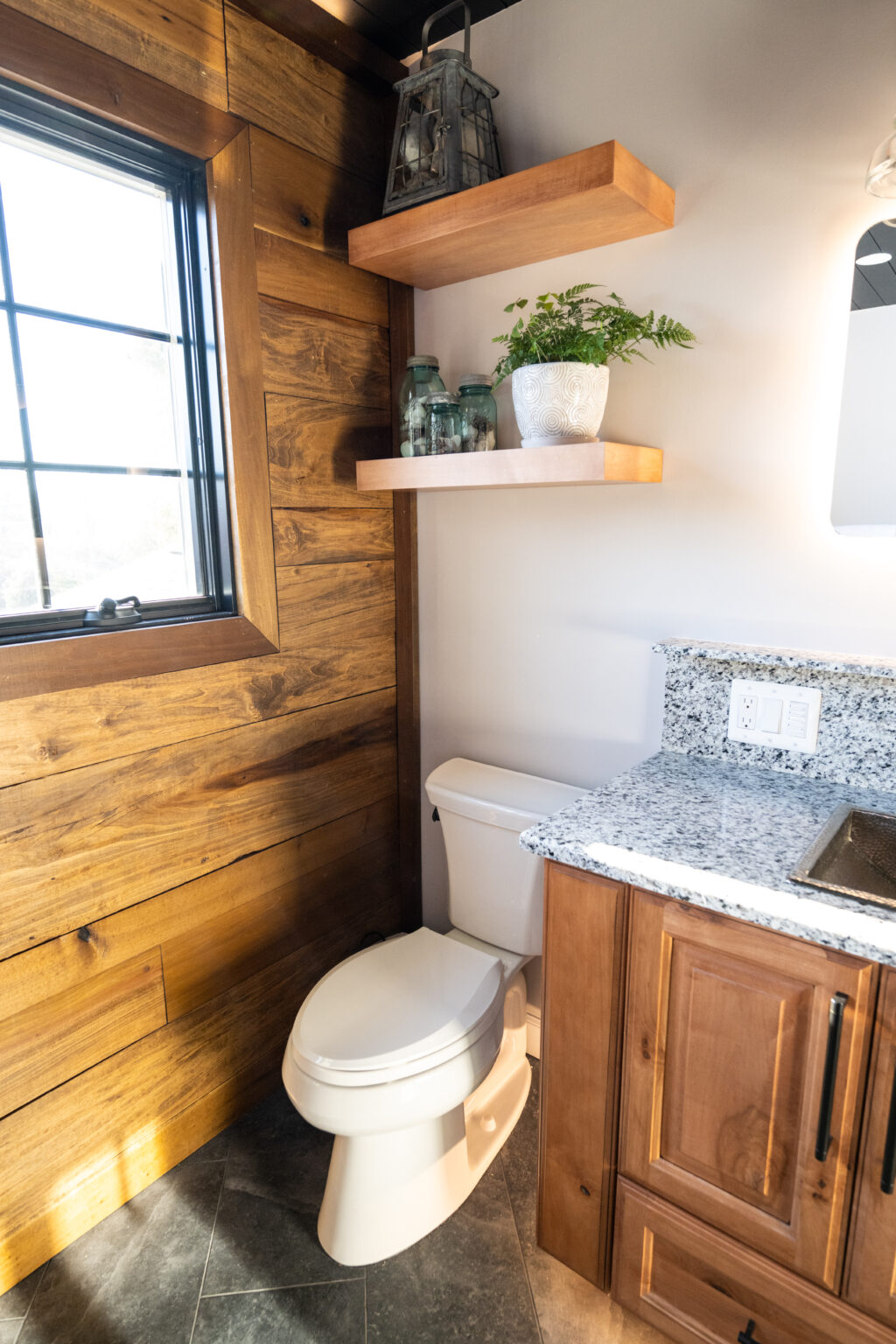 A small bathroom with a white toilet, wood-paneled walls, granite countertop, and floating shelves holding plants and jars by a window.