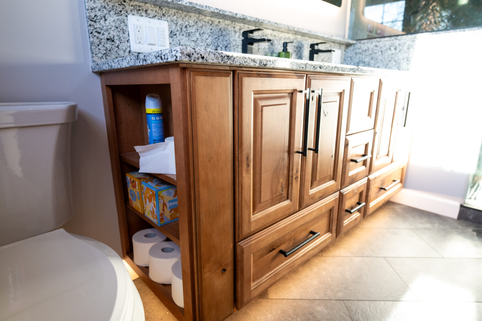 Bathroom vanity with wooden cabinets, shelves storing cleaning spray, tissues, and toilet paper rolls, next to a toilet with a granite countertop above.