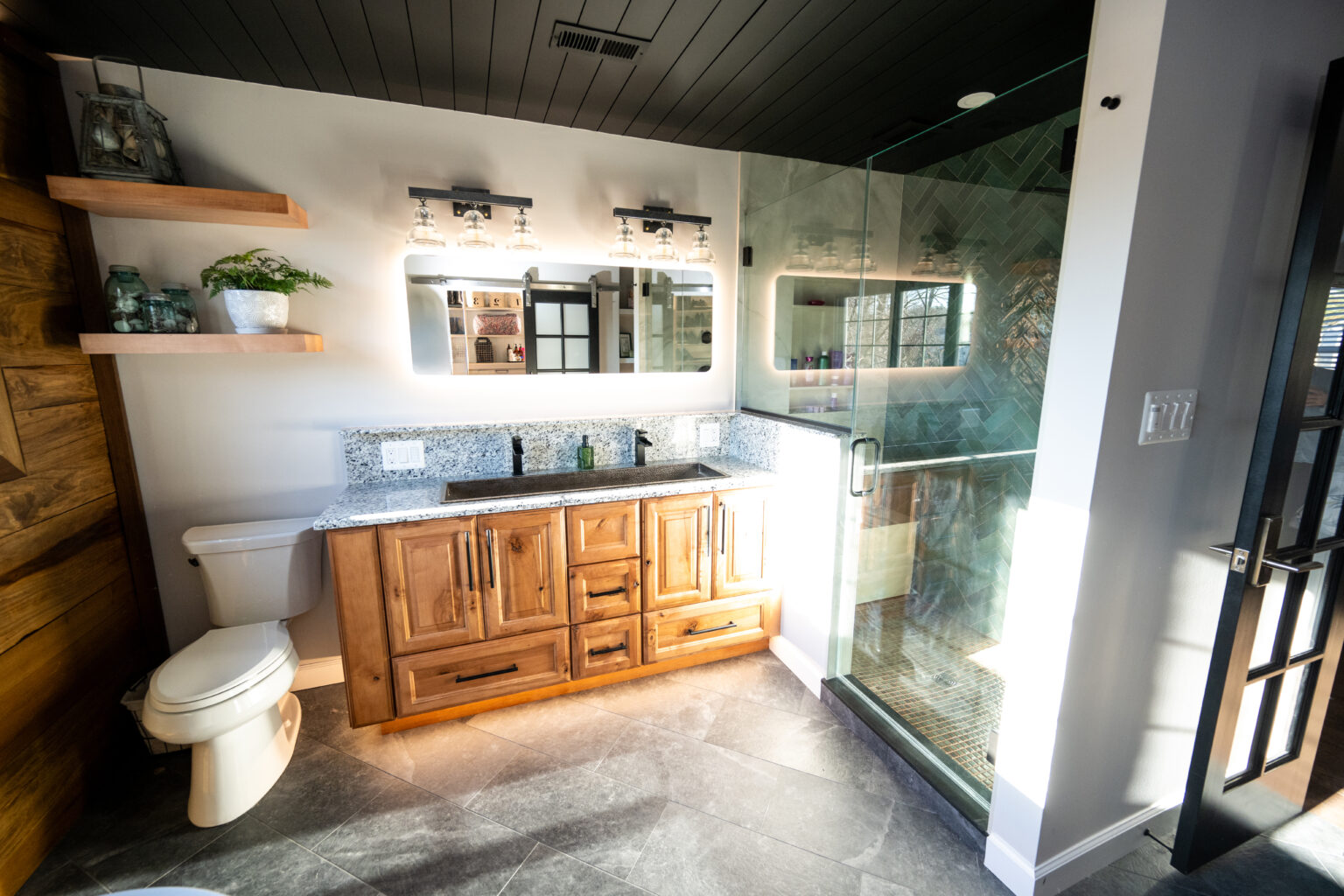 Modern bathroom with wood cabinets, granite countertop, glass shower enclosure, open shelves, wall-mounted lights, and a white toilet on gray tile flooring.