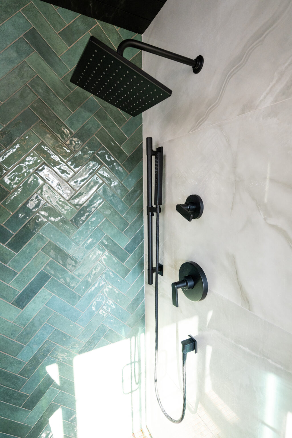 A modern shower with a rainfall showerhead, handheld shower, and matte black fixtures, set against green herringbone and white marble-look tiles.