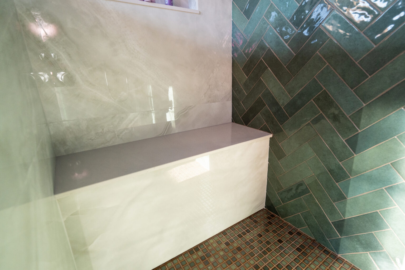 A shower features a built-in bench with a smooth white surface, beige marble-look wall tiles, green herringbone tiles, and small square brown floor tiles.
