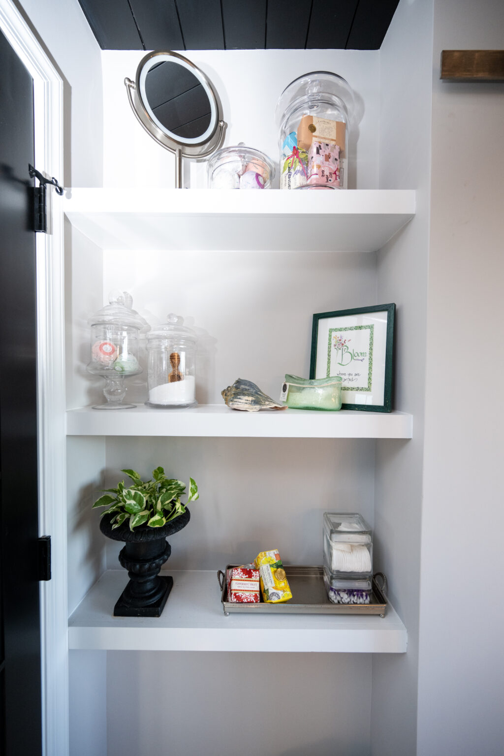Three white shelves with glass jars, a plant in a black pot, a framed picture, a makeup mirror, and a tray with soap bars and a glass container.