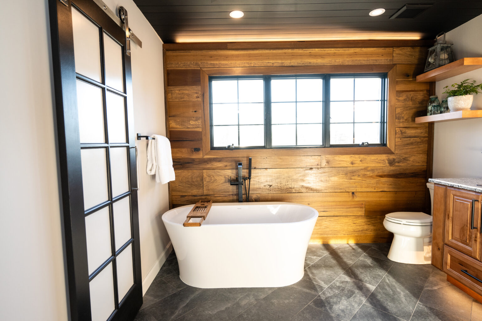Modern bathroom with a freestanding white tub, wooden accent wall, large window, towel rack, toilet, and shelves with plants and decor.