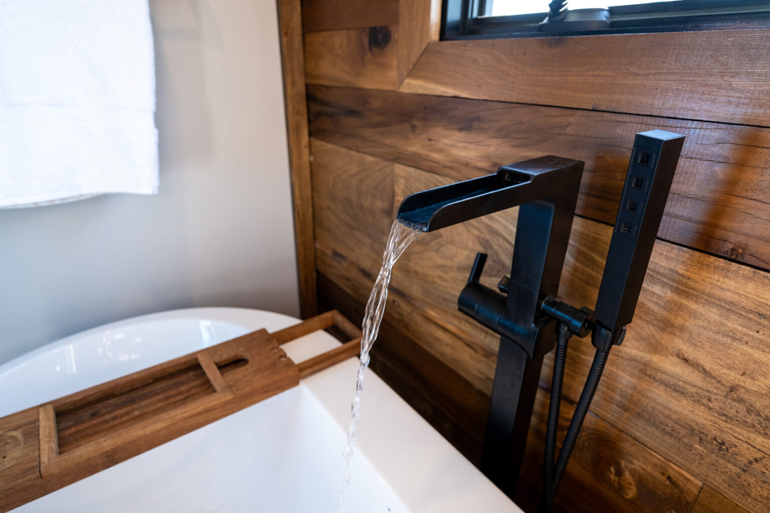 A modern black bathtub faucet pours water into a white tub, set against a wooden wall with a towel and bath caddy nearby.