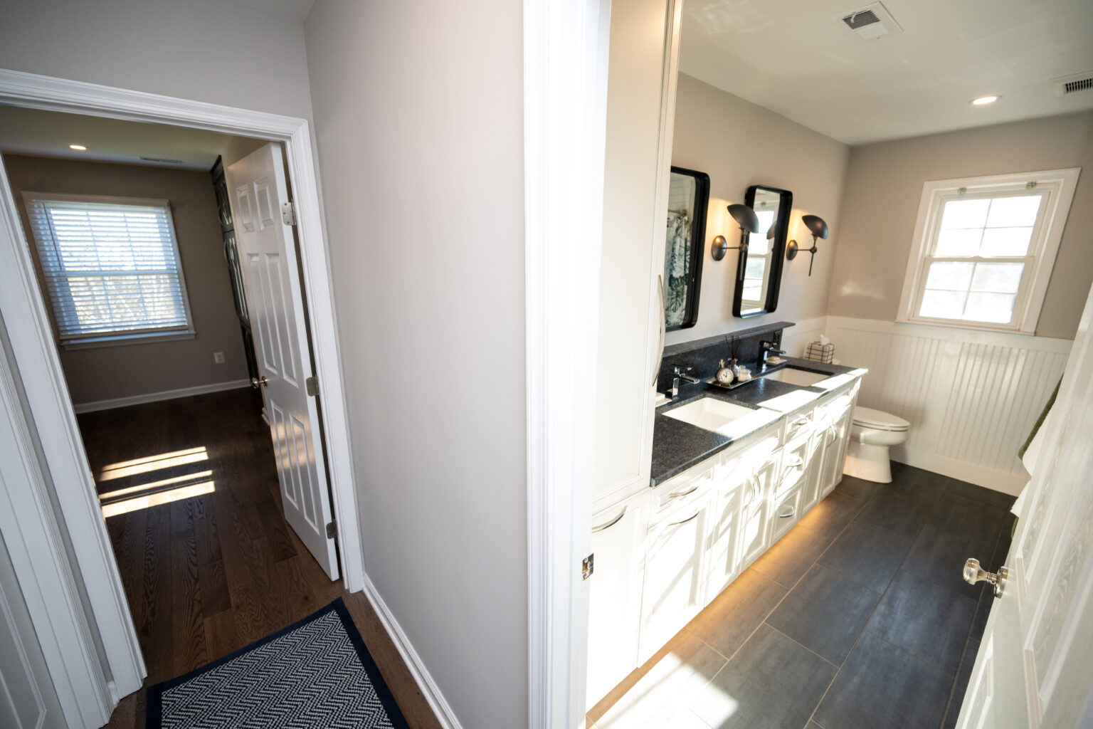 A well-lit bathroom with a double sink vanity, two mirrors, a toilet, and a window, adjacent to a hallway leading to a room with hardwood floors and a window.