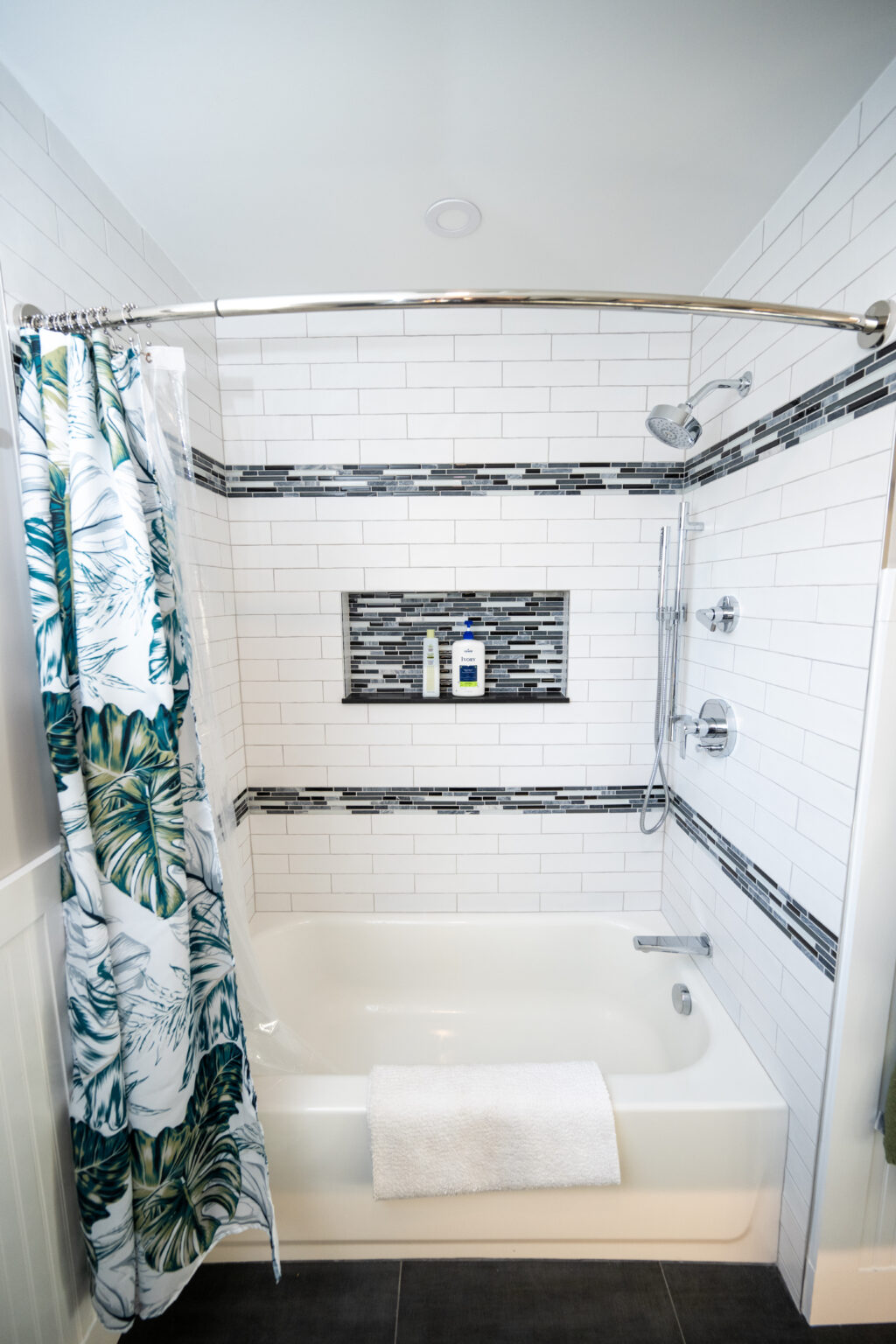 White bathroom with a bathtub, tropical-patterned shower curtain, wall-mounted shower fixtures, built-in shelf with toiletries, and a white towel on the tub edge.