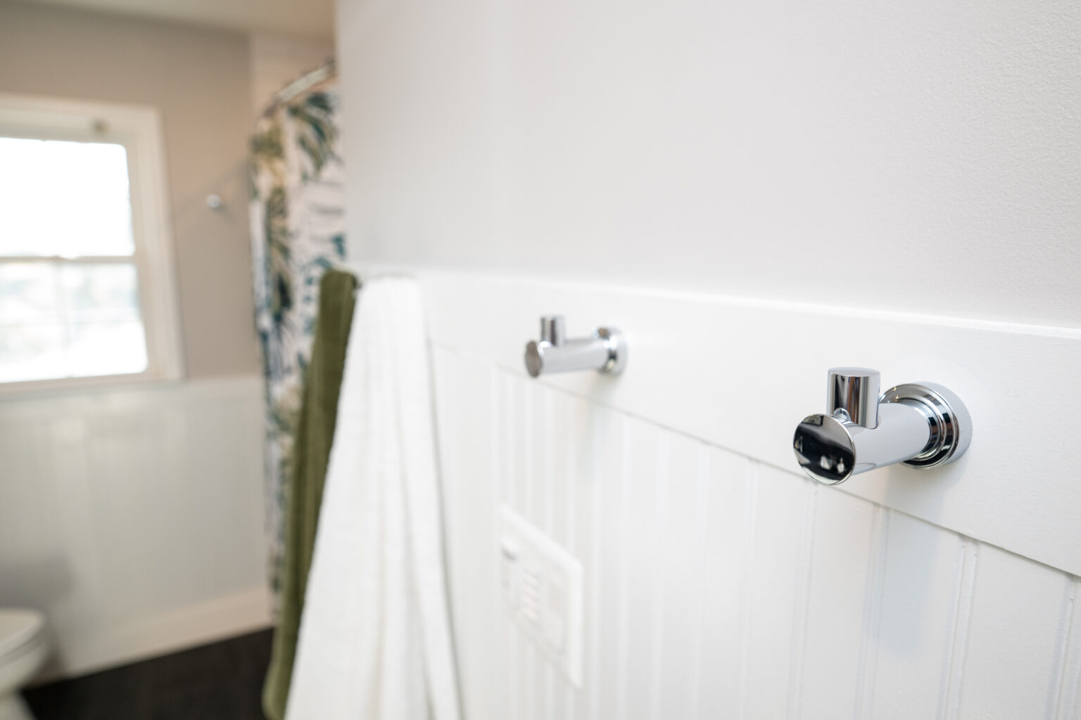 Two chrome wall hooks are mounted on white beadboard paneling in a bathroom, with towels hanging nearby and a window in the background.