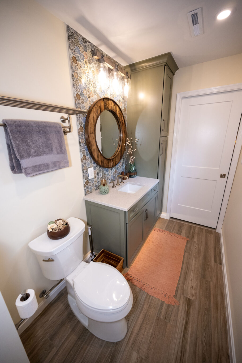 Modern bathroom with wooden flooring, a toilet, sink, circular mirror, and light fixture. Green cabinets and hexagonal tile backsplash. Tan rug is placed near the sink.