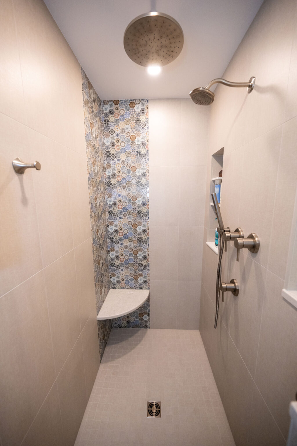 Modern shower with beige tiles, mosaic accent wall, ceiling and wall-mounted showerheads, corner shelf, and built-in niche for toiletries.